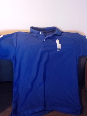 Blå pikétröja från Ralph Lauren - Klassisk blå pikétröja från Ralph Lauren med stor vit logga på bröstet och en vit siffra 3 på ärmen. Tröjan har krage, knappar vid halsen och är tillverkad i bomull. Perfekt för en sportig och stilren look. Är Large men funkar för 13 till 16 åringar