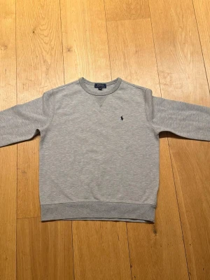Grå sweatshirt från Polo Ralph Lauren - Klassisk grå sweatshirt från Polo Ralph Lauren med rund halsringning och diskret broderad logga på bröstet. Tröjan har långa ärmar och ribbade muddar vid ärmslut och nederkant. Tillverkad i mjukt bomullsmaterial för en skön och avslappnad känsla.
