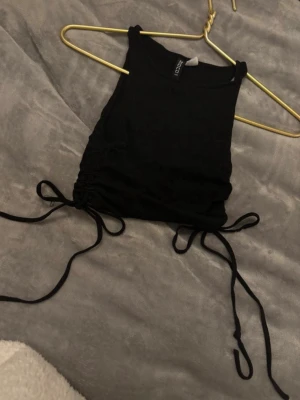 Svart croppad linnetopp från Divided - Svart croppad linnetopp från Divided med snörning i sidorna. Toppen är ärmlös och har en tight passform, perfekt att styla med höga jeans eller kjol. Materialet är ribbat och känns mjukt mot huden.