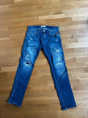 Replay jeans - Säljer ett par blå replay jeans med slitningar. Jeansen har fem fickor, normal midja och raka ben. Materialet är klassisk denim i bomull och de har en cool, avslappnad look. Perfekta för dig som gillar streetstyle och en trendig vibe. Jeansen har ett litet hål vid skrevet vilket skulle behöva fixas men det går att använda utan det.
