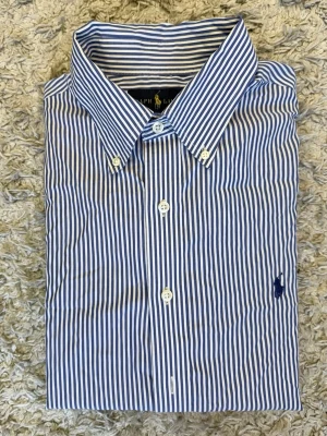 Blårandig skjorta från Ralph Lauren - Klassisk blårandig skjorta från Ralph Lauren med vit knappstängning och broderad logga på bröstet. Skjortan har lång ärm, button-down krage och är tillverkad i mjuk bomull. Perfekt för en stilren och tidlös look. Sitter som mellan M och L