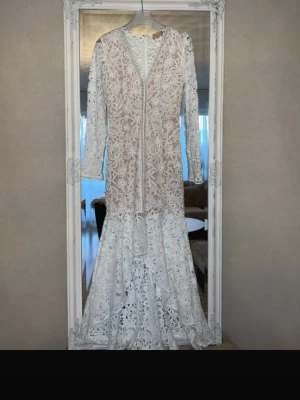 Vit lång studentklänning spetsklänning med volang - Elegant vit långklänning i spets med v-ringning och långa ärmar. Klänningen har vackra volangdetaljer nedtill och ett romantiskt blommigt mönster. Perfekt för speciella tillfällen där du vill känna dig extra fin. Nypris 2599