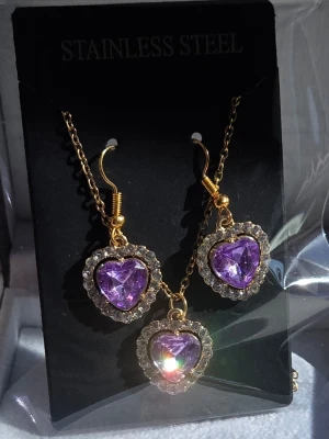 Set med lila hjärtsmycken - Stenarnas facettslipning fångar upp solens strålar och skapar ett levande ljusspel som skiftar i lila toner✨️ // Snyggt set med halsband och örhängen i guldtonad metall. Både halsbandet och örhängena har hjärtformade lila stenar omgivna av små gnistrande stenar. Materialet är rostfritt stål och designen känns både söt och trendig. Kedjan är 45 cm + förlängningskedja och hängena är ca 1,8 - 2 cm.