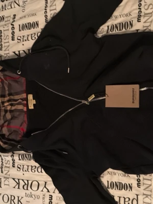 Svart hoodie från Burberry med dragkedja - Snygg svart hoodie från Burberry med dragkedja och klassiskt rutigt foder i huvan. Märket syns tydligt på etiketten och den har en justerbar huva med snören. Helt ny med prislapp dessutom! 