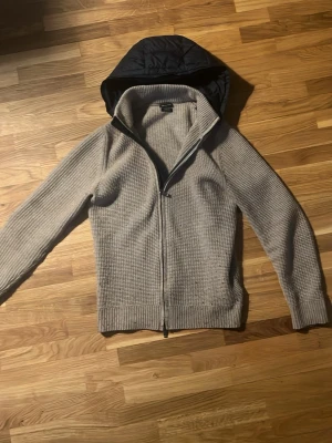 Massimo dutti cardigan  - Säljer en beige stickad hoodie med dragkedja framtill och svart quiltad huva. Tröjan har ribbade muddar vid ärmslut och nederkant samt en avslappnad passform. Perfekt för dig som gillar en casual och stilren look.