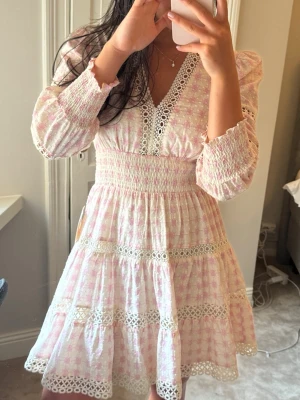 Inez dress. Rosa och vit by malina klänning med spets - Supersöt klänning i rosa och vitt. V-ringad med spetsdetaljer längs urringning och kjol, smockad midja och långa ärmar med smock vid ärmslut. 