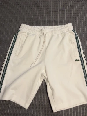 Vita shorts från Lacoste med snörning - Säljer ett par vita shorts från Lacoste med gröna ränder längs sidorna och klassisk krokodillogo. Shortsen har elastisk midja med snörning och fickor fram samt en bakficka. Perfekta för sommaren och sportiga outfits. Skicka meddelande vid frågor.