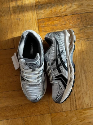 Asics gel kayano 14 JJJJOUND - Snygga sneakers från ASICS i silver och vitt med svarta detaljer. Ovandelen är i mesh och syntet för bra andningsförmåga och komfort. Klassisk låg modell med tydliga ASICS-loggor på sidorna och robust sula i svart och vitt. Perfekta för dig som gillar sportig stil.