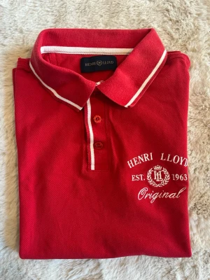 Pikétröja från Henri Lloyd - Klassisk röd pikétröja från Henri Lloyd med vit brodyr på bröstet och vita detaljer på krage och knappslå. 