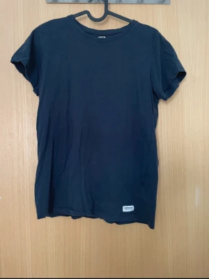 Svart basic t-shirt från Lager 157 - En svart basic t-shirt från Lager 157 med rund hals och korta ärmar. Tillverkad i mjuk bomull och har en liten diskret logotyp-patch nertill. Perfekt till jeans eller shorts för en clean look.