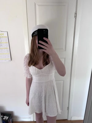 Vit spetsklänning med puffärm - Söt vit kortklänning med spetsdetaljer och puffiga kortärmar. Klänningen har v-ringad urringning och markerad midja som ger en feminin siluett. Perfekt till student eller sommarens höjdpunkter.