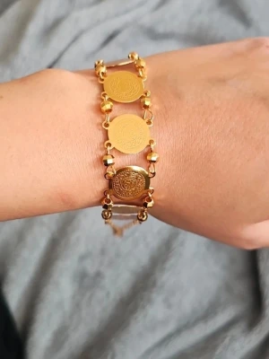 Guldigt armband med myntdetaljer - Snyggt armband i guldfärgad metall med flera runda myntliknande plattor och små pärlliknande detaljer mellan länkarna. Armbandet har en kedja och stängs med ett klassiskt lås. Perfekt accessoar för att lyfta din stil.