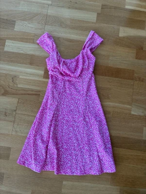 Rosa blommig kortklänning - Söt rosa klänning med vita små blommor och breda axelband. Klänningen har en lätt utsvängd kjol och mjukt fallande tyg, perfekt för varma dagar. 