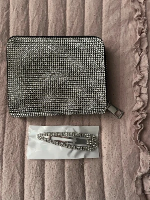 Glittrig plånbok med strassdetaljer - Säljer en fyrkantig plånbok täckt av glittrande strass i silverfärg. Plånboken har dragkedja runtom och flera fack inuti för kort och sedlar. Perfekt accessoar för dig som gillar att sticka ut med lite extra bling.