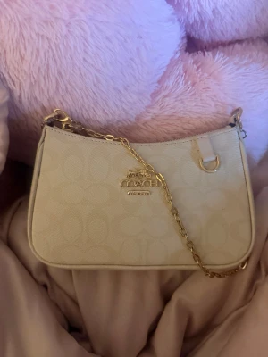 Beige axelremsväska från Coach - Snygg beige handväska från Coach med guldfärgad kedja och diskret logomönster över hela väskan. Väskan har en stilren rektangulär form och detaljer i guld, samt Coach-logga framtill. Perfekt för dig som gillar klassisk och trendig design.