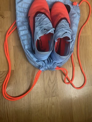 Nike Phantom Elite fotbollsskor storlek 43 - Säljer ett par Nike Phantom Vision fotbollsskor i en snygg kombination av ljusblått och neonorange med svarta detaljer. Skorna har snörning och är designade för bra bollkontroll och grepp på planen. Perfekta för dig som vill sticka ut på matchen. Använda väldigt få gånger, pris kan diskuteras vid snabb affär.