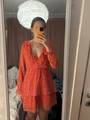 Röd blommig omlottklänning H&M - Supersöt röd omlottklänning från H&M med små vita blommor, volangdetaljer vid halsringningen och långa ärmar med resår vid handlederna. Klänningen har markerad midja och kjol med volangkant. Perfekt för vår och sommar! Knappen uppe har dock gått sönder, så om du är duktig på att sy är det lätt att lösa om du vill ha den mindre urringad!