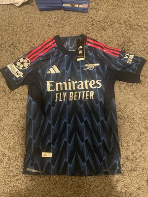 Arsenal gyökeres adidas bortatröja - Snygg Arsenal bortatröja från Adidas med Gyökeres och nummer 14 på ryggen. Tröjan är mörkblå med ljusblå blixtmönster, röda ränder på axlarna och vita loggor. Tillverkad i lätt och ventilerande material, perfekt för fotboll eller träning. Den är  aldrig använd. Kommer i original adidas kasse. Pris kan diskuteras