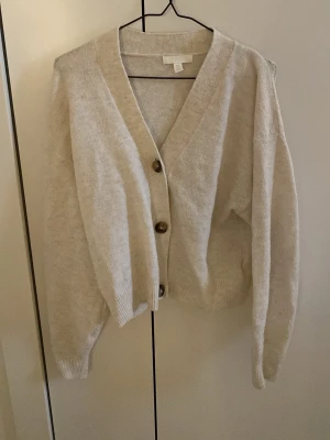 Beige kofta från H&M med knappar - Mysig beige kofta från H&M med stora bruna knappar framtill. Koftan har v-ringning, ribbade muddar och en avslappnad passform. Perfekt att slänga över en t-shirt eller topp för en chill look.