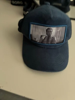 Mörkblå keps från Contore - Snygg mörkblå keps från Contore med unik meshdetalj framtill som visar ett svartvitt foto. Kepsen har klassisk böjd skärm och justerbar baksida. Insidan har meshfoder och band med Contore-logga. Perfekt för dig som vill sticka ut med din stil.