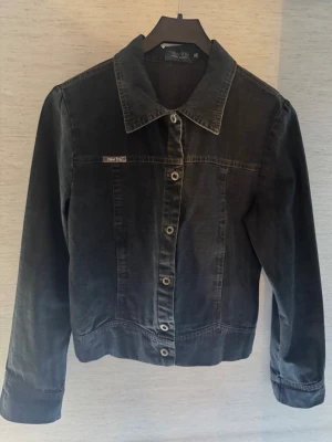 Svart jeansjacka från New Line - Snygg svart jeansjacka från New Line med klassisk krage och raka ärmar. Jackan har knäppning framtill med metallknappar och diskret logga på bröstet. Perfekt för dig som gillar en stilren och tidlös look.