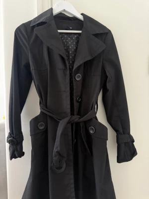 Svart trenchcoat med bälte och knappar - Stilren svart trenchcoat med klassisk krage, stora knappar och knytskärp i midjan. Jackan har långa ärmar med knäppning vid ärmslut och fickor framtill. Perfekt för dig som vill ha en tidlös och snygg look.