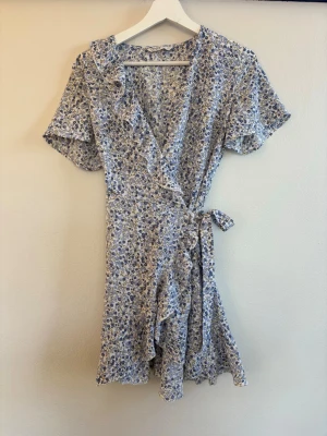 Blommig omlottklänning från ONLY - Söt kort omlottklänning från ONLY med blått och vitt blommönster. Klänningen har volangdetaljer, knyt i midjan och korta ärmar. Perfekt för sommarens alla tillfällen.