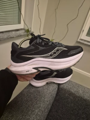 Svarta Saucony Axon 2 sneakers med vit sula 37.5 - Säljer ett par svarta Saucony sneakers med vit tjock sula och silverfärgade detaljer på sidan. Skorna har mesh-överdel för bra ventilation och klassisk snörning. Perfekta för dig som gillar sportig stil och vill ha bekväma skor till vardags.