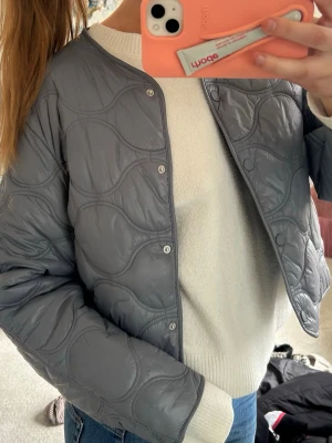 Grå quiltad cardiganjacka - Snygg grå quiltad cardiganjacka med rund halsringning och tryckknappar framtill. Jackan har ett vågigt mönster i sömmarna och är tillverkad i ett glansigt material som ger en modern look. Perfekt att slänga över en tröja för extra stil. Passar ca M❤️