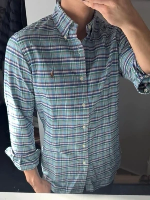 Rutig skjorta från Ralph Lauren - Säljer en klassisk rutig skjorta från Ralph Lauren i blå, grön och vit färg. Skjortan har lång ärm, knappar framtill och button-down krage. Tillverkad i mjuk bomull och har den ikoniska broderade loggan på bröstet. Perfekt för en stilren och avslappnad look.