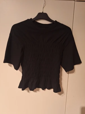 Svart topp från Gina Tricot - Svart topp från Gina Tricot med smockad midja och lätt utsvängd nederkant. Toppen har korta ärmar och rund halsringning. Perfekt att styla med både jeans och kjol för en trendig look.