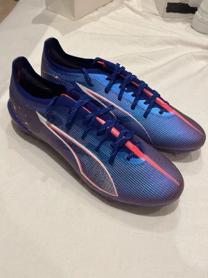 Blåa Puma fotbollsskor med röd detalj - Säljer ett par snygga blåa fotbollsskor från Puma med röda och vita detaljer. Skorna har låg profil, snörning och en lätt, syntetisk ovandel som ger bra bollkontroll. Perfekta för dig som vill sticka ut på planen och få grym känsla i spelet.