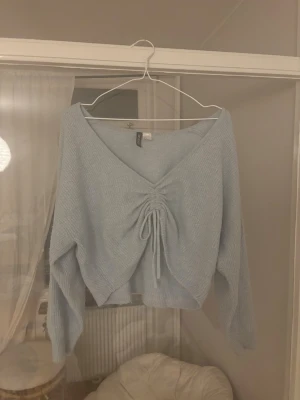 Ljusblå croppad topp från H&M Divided - Supersöt ljusblå croppad topp från H&M Divided med v-ringning och snörning framtill. Toppen har långa ärmar och är gjord i ett mjukt, ribbat material som känns skönt mot huden. Perfekt för dig som gillar trendiga och bekväma plagg. Tyvärr är tröjan ganska nopprig.