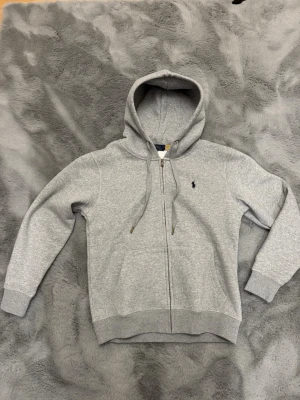 Grå hoodie från Polo Ralph Lauren - Snygg grå hoodie från Polo Ralph Lauren med dragkedja framtill och klassisk huva med dragsko. Liten broderad logga på bröstet och ribbade muddar vid ärmar och midja. Tillverkad i mjuk bomullsblandning för extra komfort.