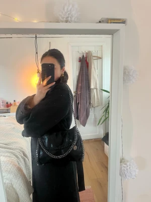 Svart handväska med kedjedetaljer -  svart handväska med glansig finish och kraftiga kedjor längs kanterna och som axelband. Väskan har en flap-öppning och dekorativ söm runtom.