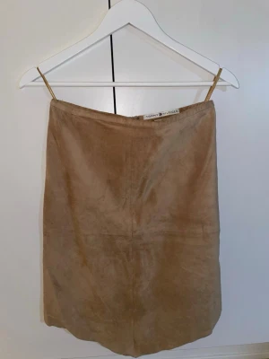 Beige mockakjol från Tommy Hilfiger - Snygg beige knappt knäkjol från Tommy Hilfiger i mjuk mocka. Kjolen har enkel design med snedskurning nedtill och dragkedja bak. Perfekt för dig som gillar stilrena plagg med en lyxig känsla. Aldrig använd. 