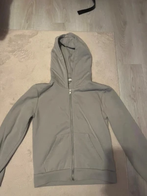 Grå zip hoodie med fickor - En stilren grå hoodie med dragkedja framtill och två stora fickor. Hoodien har en klassisk huva och ribbade muddar vid ärmslut och nederkant. Perfekt för en avslappnad look och enkel att matcha med andra plagg.