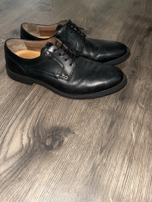 Svarta klassiska loafers i skinn - Stilrena svarta loafers i äkta skinn med rundad tå och diskreta sömmar. Skorna har snörning framtill och en låg klack. Insidan är ljusbrun och har en präglad läderdetalj. Perfekta för dig som gillar en clean och tidlös look.
