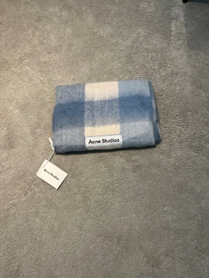 Blårutig halsduk från Acne Studios - Supersnygg halsduk från Acne Studios i mjuk ullblandning. Den har ett rutigt mönster i ljusblått, vitt och beige samt en tydlig Acne Studios-logga. Perfekt accessoar för att lyfta din outfit och hålla dig varm under kalla dagar.