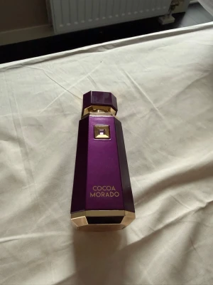 French Avenue Cocoa Morado parfym  - Lyxig parfymflaska från Cocoa Morado i en djup lila färg med eleganta gulddetaljer. Flaskan har en geometrisk, modern form och känns exklusiv med sin metalliska finish. Perfekt för dig som gillar unika och snygga parfymflaskor.
