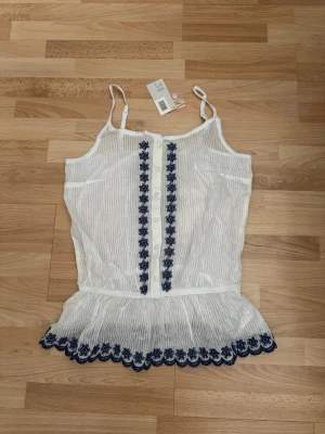 Ny. Vit blommig linnetopp med spets - Söt vit linnetopp med tunna axelband och blå broderade blommor längs framsidan och nederkanten. Toppen har volang nertill och är tillverkad i ett lätt, randigt bomullstyg. Perfekt för en fräsch och somrig look.