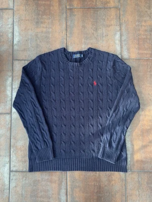 Kabelstickad Tröja Polo Ralph Lauren - Kabelstickad mörkblå tröja från Polo Ralph Lauren med rund halsringning och röd broderad logga på bröstet. Tröjan har långa ärmar och ribbade muddar vid ärmslut och nederkant. Perfekt för dig som gillar klassisk och stilren design. 