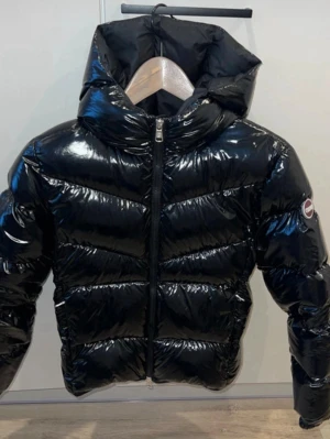 Svart glansig pufferjacka  - Snygg svart pufferjacka från COLMAR med glansig finish och stor huva. Jackan har dragkedja framtill och COLMAR-logga på ärmen. Perfekt för kalla vinterdagar och ger en riktigt trendig look.