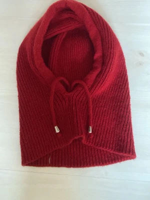 Röd stickad balaclava med snören - Säljer en ribbstickad balaclava i rött med dragsko och metalländar på snörena. Mjuk och varm, perfekt för kalla dagar. Ingen synlig logga eller märke. Materialet ser ut att vara ull eller en ullblandning.