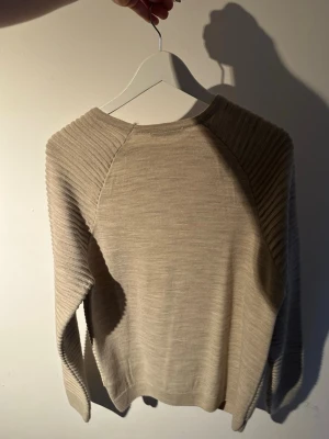 Beige ribbad stickad tröja - Säljer en beige stickad tröja med ribbad struktur och rund hals. Tröjan har långa ärmar och en avslappnad passform. Perfekt för dig som gillar stilrena och enkla plagg med lite textur. Passar till det mesta och är superenkel att matcha.