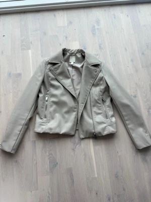 Beige skinnjacka från H&M - Snygg beige skinnjacka från H&M i klassisk bikerstil med bred krage och dragkedjor på sidorna. Jackan har en kortare passform och detaljerade sömmar som ger en cool look. Perfekt att slänga över en hoodie eller t-shirt för en trendig outfit.