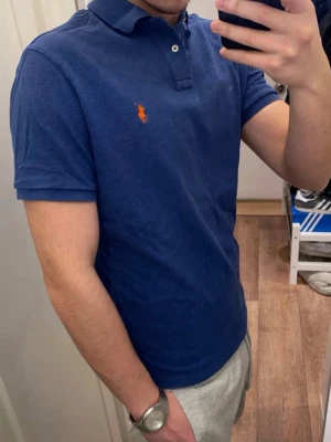 Polo Ralph Lauren (Blå piké) - Snygg blå piké från Polo Ralph Lauren med klassisk krage och knappar framtill. Ikonisk röd broderad logga på bröstet. Den är oanvänd och redo för en ny ägare! Pikén är storlek L men passar även M!