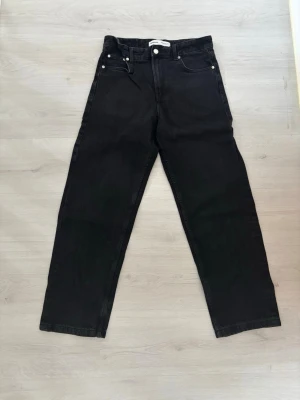 Svarta raka jeans från Bershka - Säljer ett par svarta jeans från Bershka med rak passform och klassisk femficksdesign. Jeansen har hög midja, bälteshällor och silverfärgade knappar. Materialet är denim i bomull. Perfekta för en avslappnad och trendig look.