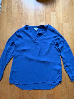 Kornblå v-ringad blus - En stilren kornblå blus med v-ringning och långa ärmar. Blusen har en avslappnad passform och rundad nederkant. Tillverkad i ett mjukt, lätt material som ger ett snyggt fall. Perfekt att matcha med jeans eller kjol för en trendig look.