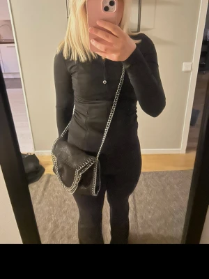 Svart Stella McCartney liknande handväska - Säljer en svart handväska som liknar Stella McCartney med kedjedetaljer längs kanterna och axelrem i silverfärgad metall. Väskan har en mjuk, glansig yta och en avslappnad, fyrkantig form. Perfekt för dig som gillar trendiga accessoarer med coola detaljer.